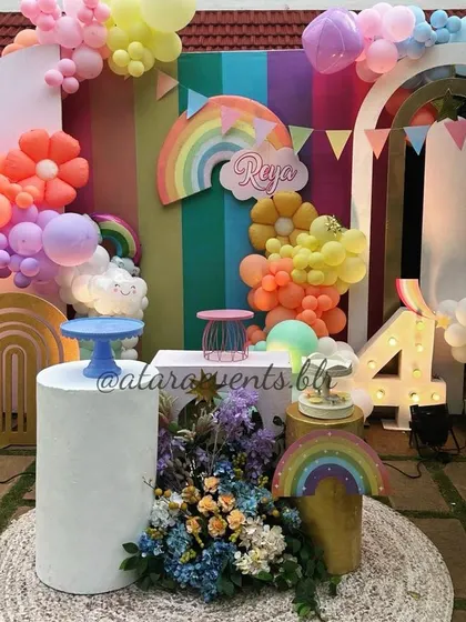 A vibrant rainbow-themed birthday party, bursting with color and joy. The decor features a multi-colored striped backdrop, cheerful rainbow and cloud cutouts, and a '4' marquee light, making for a picture-perfect celebration.