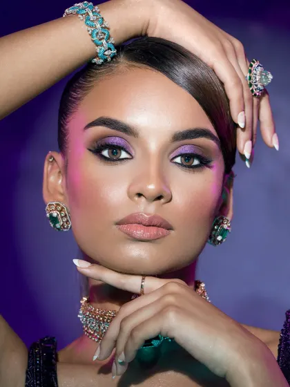 A dramatic beauty shot with vibrant purple eyeshadow and expertly placed highlights. The pose draws attention to the stunning emerald jewelry and flawless manicure.