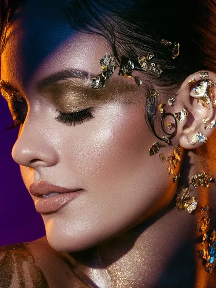 A macro detail of the gold foil makeup. This close-up reveals the intricate application of the gold leaf on the skin, hair, and ear, showcasing the artistry and texture of this avant-garde look.