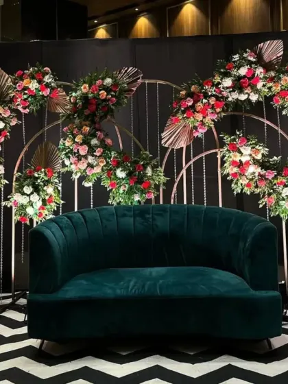 A glamorous party decor setup with a deep green velvet sofa. The backdrop features metal arches adorned with beautiful red and pink floral arrangements.