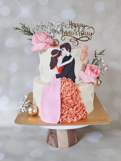 A romantic two-tier anniversary cake featuring a hand-painted couple illustration. The bride's ruffled dress is created with delicate buttercream piping, adding a three-dimensional, artistic touch to the design.