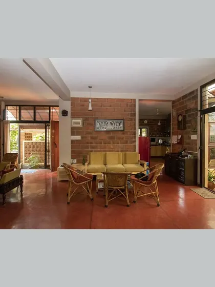 The main living area, designed as a modern 'mandapa', with cane furniture, red oxide flooring, and sliding doors opening to a garden.