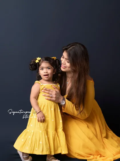 A beautiful mother-daughter portrait in matching yellow dresses against a classic dark backdrop. The daughter's joyful expression says it all.