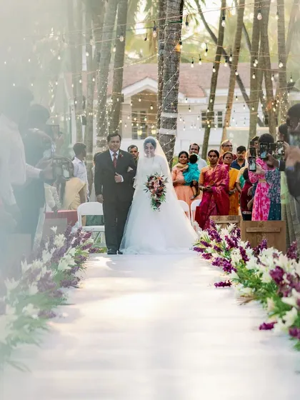 A bride's unforgettable walk down an aisle lined with purple and white flowers. The hanging lights add a magical touch to this outdoor Christian wedding ceremony at Adyar Garden.