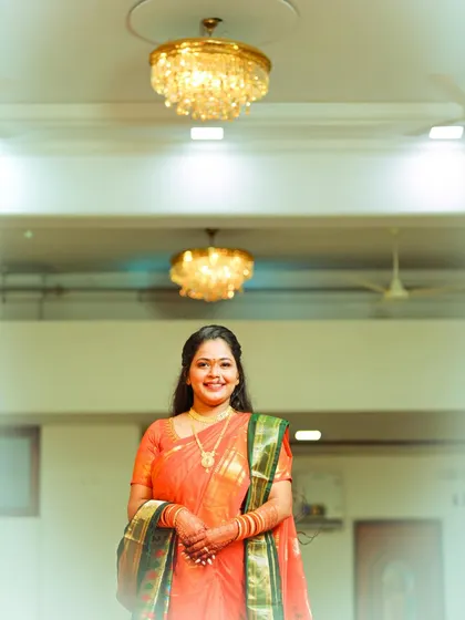 A lovely solo portrait of the bride in her orange saree, standing under a chandelier. The soft, bright lighting gives the image an ethereal and celebratory feel.