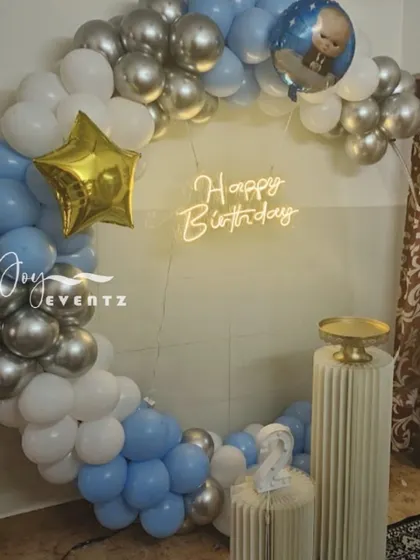 This is another take on the balloon ring decoration, perfect for a home setting. The arch of blue, white, and silver balloons is accented with a star foil balloon and a 'Happy Birthday' neon sign, with a small Boss Baby balloon detail.