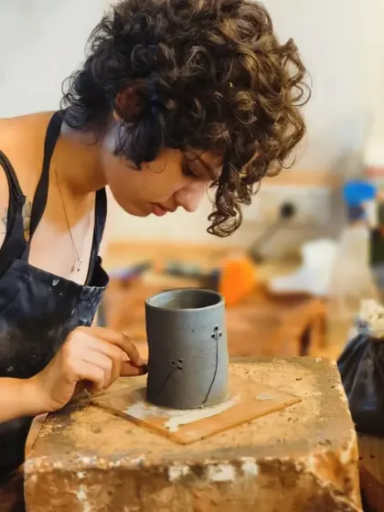 A student carefully carves delicate dandelion patterns onto her grey stoneware cup. This sgraffito technique allows for fine, detailed illustrations on clay.