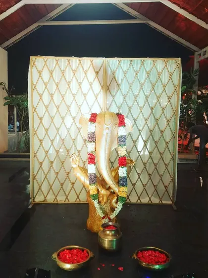 This image shows the Ganesha idol with a traditional garland against a diamond-patterned backdrop. Small bowls of flower petals are placed in front for offerings.
