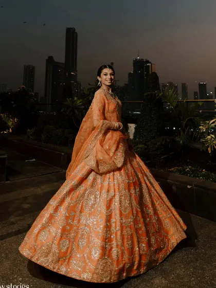A breathtaking wide shot of the bride in her magnificent orange lehenga for her pre-wedding reception. The makeup is elegant and defined to balance the grandeur of the outfit.