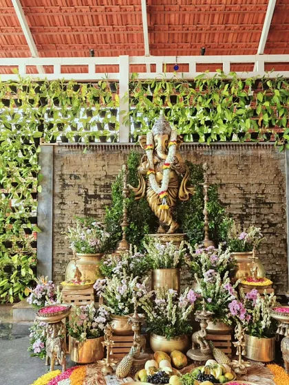 A lush and divine Ganesha setup for a blessed beginning. The idol is surrounded by an abundance of fresh flowers, fruits, and traditional brass pots against a vertical garden wall.