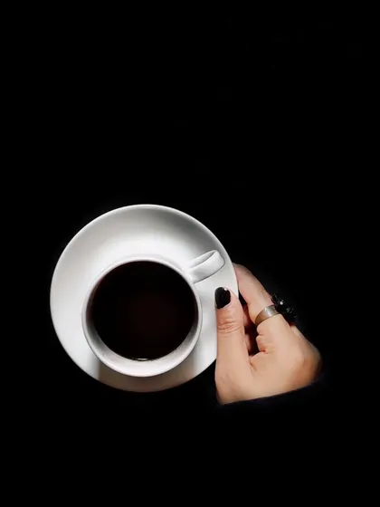 A minimalist, high-contrast shot of a hand holding a white cup of black coffee against a pure black background. This is a powerful, graphic image that speaks of simplicity and elegance.