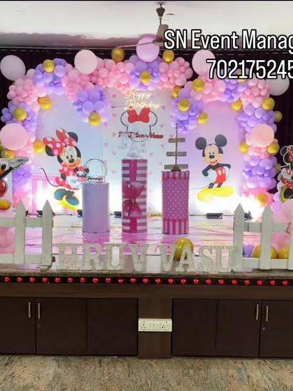 A cute and classic Minnie and Mickey Mouse stage decoration. The setup includes a picket fence, character cutouts, and a pink and purple balloon arch, perfect for a toddler's birthday.