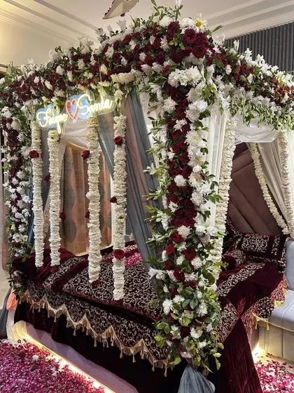 A close-up of a luxurious wedding bed decoration. The four-poster frame is heavily decorated with red and white flowers and hanging floral strings, creating a stunning floral cocoon.
