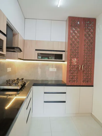This L-shaped kitchen features a unique tall pantry unit with decorative CNC-cut doors, adding a traditional touch to the modern layout. The combination of light wood and white cabinets keeps the space feeling bright and airy.