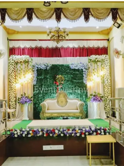 A full view of a stage setup within a conventional hall, showing how we transform a space. This design includes a green wall, elegant seating, and floral arrangements, framed by the hall's own traditional curtains.