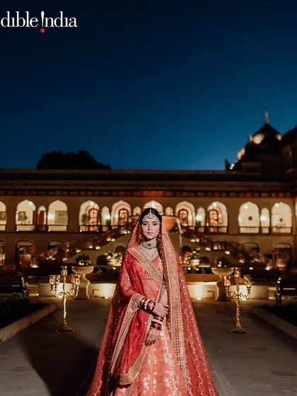 A bride stands in her red wedding attire against the illuminated facade of Rambagh Palace at night, a vision of timeless elegance.