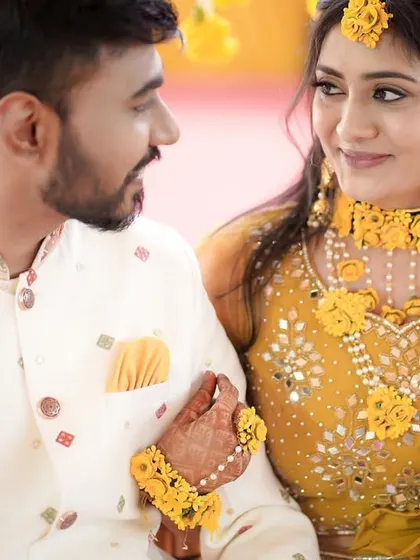 A sweet moment between the couple at their Haldi function. The bride holds onto the groom's arm as they share a look, surrounded by traditional yellow decor.