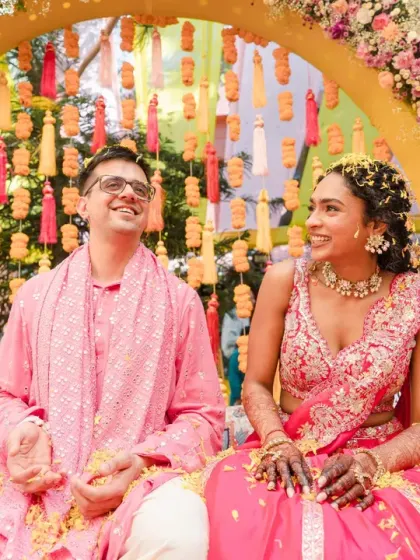 The couple sits together, smiling, under a vibrant floral arch. This shot captures a moment of shared happiness before the main haldi chaos begins.