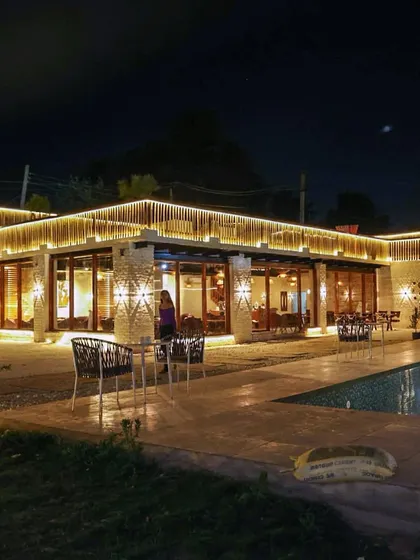 A full view of the cafe building from the poolside at night. The warm lights from inside create a welcoming glow.