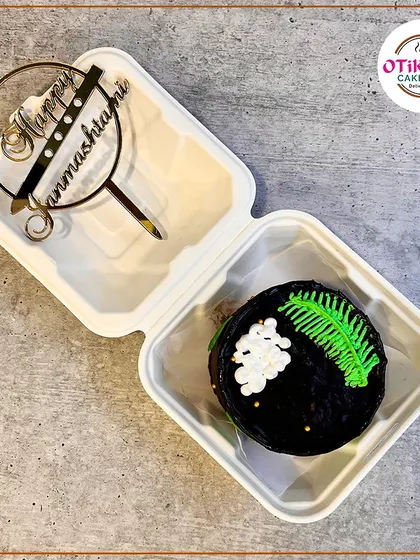 A modern and chic Janmashtami bento cake. The dark chocolate base is decorated with a delicate, hand-piped green leaf and a custom "Happy Janmashtami" topper.