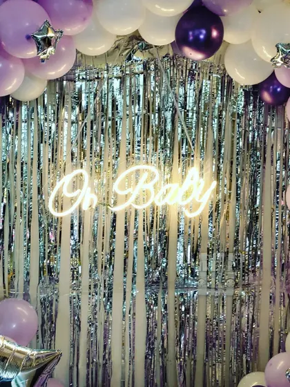 The centerpiece of this baby shower decor is the "Oh Baby" neon sign against a sparkling silver curtain, framed by a lush arch of lavender, purple, and white balloons.