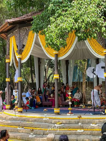 A traditional wedding ceremony in progress at a beautiful heritage venue. The mandap is adorned with yellow and white floral drapes.