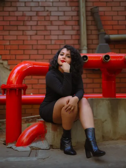 A seated portrait in an industrial setting. The contrast between the model's black dress and the bright red pipes creates a visually striking composition.