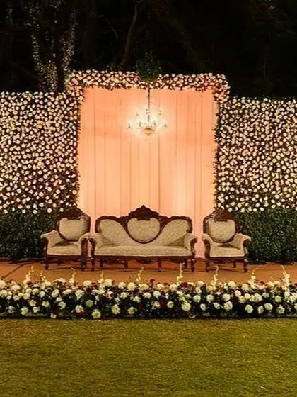 A beautiful Sangeet stage featuring a massive wall of white and red flowers. The vintage-style seating and a central chandelier add a touch of classic elegance to the outdoor lawn setting.