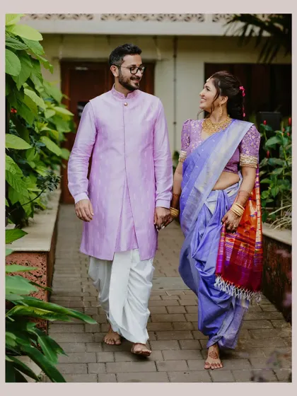 A Maharashtrian couple walks hand-in-hand through a lush green pathway, sharing a look of love and companionship in their traditional wedding attire.