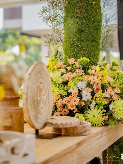 A close-up of the decor from our 'Golden Origami Garden' theme. A carved wooden artifact stands beside a lush arrangement of yellow and peach flowers, evoking a sense of warmth and artistry.