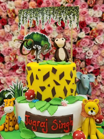 A two-tiered jungle theme cake for a first birthday. It's covered in fondant animals like a lion, giraffe, and monkey, with a fun giraffe-print pattern on the top tier.