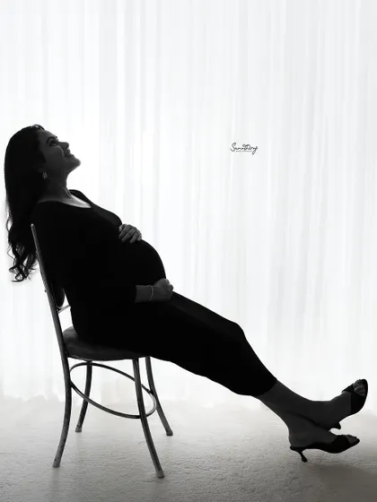 A serene and contemplative silhouette of a mother-to-be seated by a window. The bright backlighting creates a peaceful and artistic portrait that emphasizes her beautiful form.