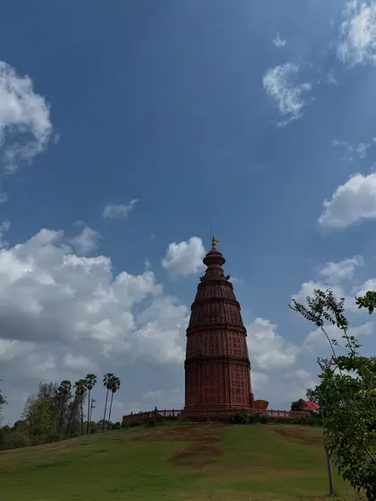 A tall, red-brick temple tower at Govardhan Ecovillage, standing against a bright blue sky with fluffy clouds.
