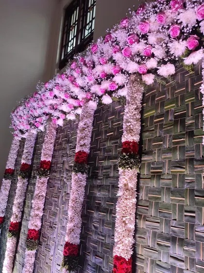 This decor features thick, beautiful flower pillars made of white and red blooms against a rustic backdrop of woven palm leaves. This style is perfect for creating a rich, traditional atmosphere for any pooja or home ritual.