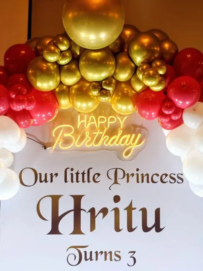 A personalized birthday backdrop for 'Our little Princess', featuring a modern white panel with custom text, framed by a red, gold, and white balloon arch and a neon sign.