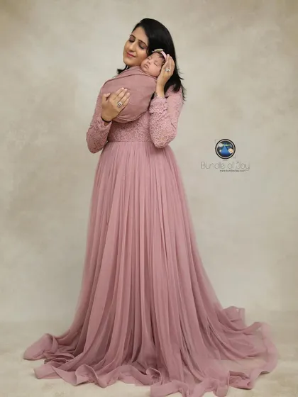 A full-length portrait of a mother in a flowing pink gown, holding her swaddled newborn close to her heart. This is a classic and graceful mother-child portrait.