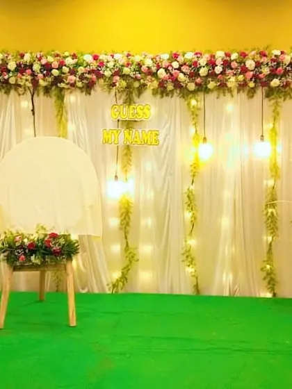 A simple and sweet naming ceremony backdrop with a "Guess My Name" sign. The white drapes, fairy lights, and floral arrangements create a warm and inviting atmosphere.