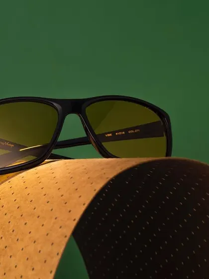 A sophisticated shot of eyewear resting on a curved piece of textured paper. The contrast between the green background and the brown paper creates a rich, earthy color palette.