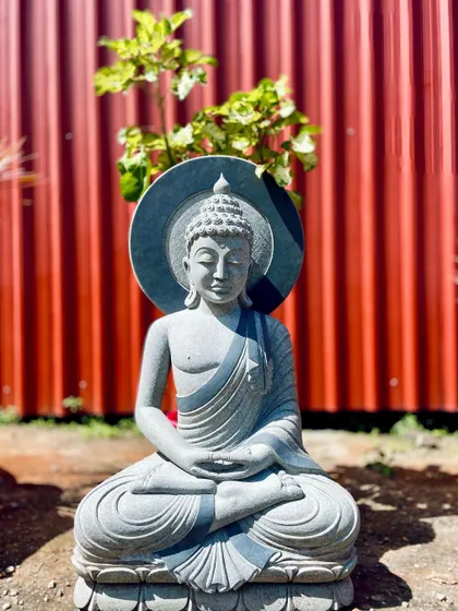 A stone Buddha statue with a halo, placed in front of a red wall. The sunlight highlights the calm expression of the sculpture.