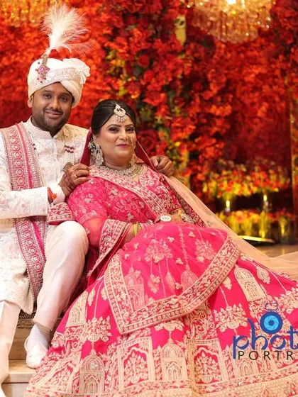 A regal portrait of the bride and groom seated against a stunning red floral backdrop, showcasing the grandeur of the decor.