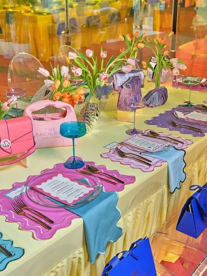 A whimsical and colorful brunch tablescape designed for a couture event. The setting features playful placemats, vibrant glassware, and handbags integrated into the decor, blurring the lines between fashion accessories and table art.