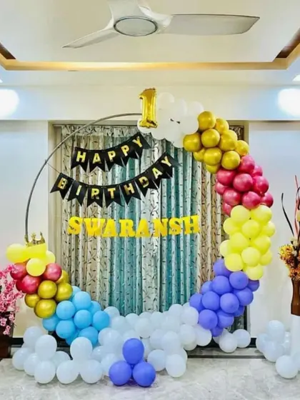 A colorful ring decoration perfect for a living room. This setup for Swaransh's first birthday uses a circular frame decorated with a rainbow of balloons and a "Happy Birthday" banner.