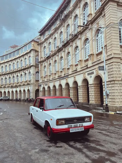A classic Premier Padmini, with its distinctive red and white paint, parked in front of the beautiful architecture of Horniman Circle.
