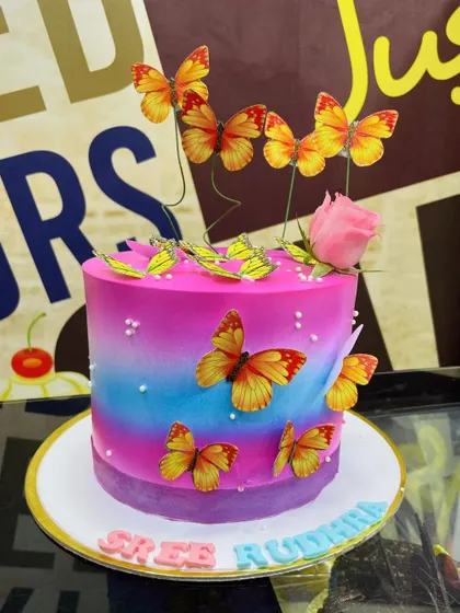 A vibrant 2kg cake with a pink, blue, and purple gradient, covered in beautiful orange and yellow butterflies.