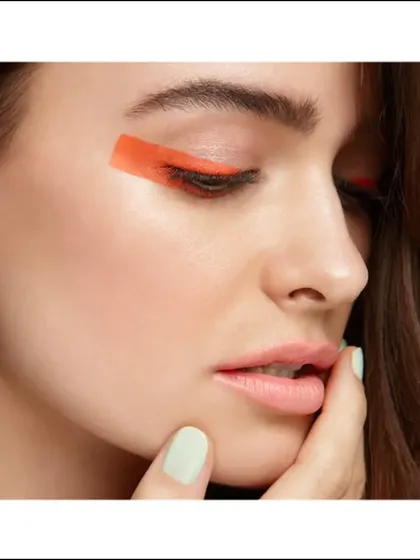 An editorial beauty look featuring a graphic swipe of orange eyeliner. The image is clean and modern, highlighting a creative and artistic approach to makeup.