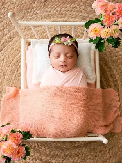 A sleeping newborn is tucked into a miniature white bed with a peach blanket, showcasing a sweet and classic prop setup for newborn photography.