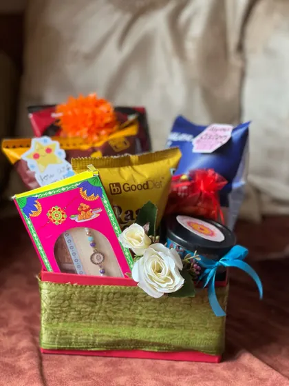 A compact Rakhi hamper in a fabric-covered box, filled with snacks, a candle, and a rakhi, adorned with small white roses.