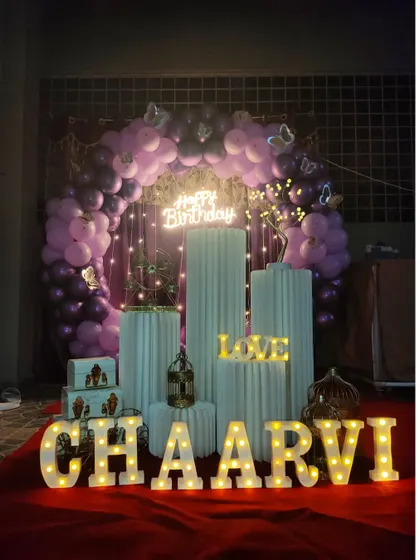 A full view of the purple butterfly-themed birthday setup, featuring a balloon ring, white cylindrical stands, and marquee letters.
