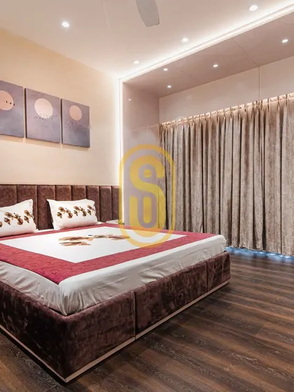 A bedroom featuring a rich, dark wood floor and a plush, channel-tufted velvet headboard. The indirect lighting from the false ceiling and beneath the curtains creates a warm and relaxing mood.