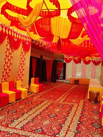 Inside a large tent set up for a housewarming. The ceiling is heavily decorated with red, yellow, and pink drapes, and the seating is arranged neatly along the carpeted floor.